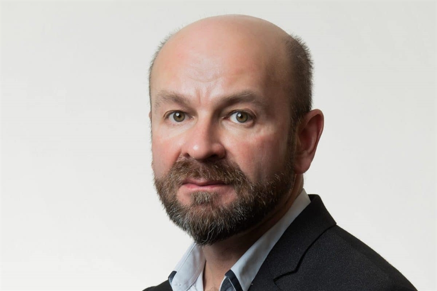 [ai] A middle-aged man with a bald head and a short beard, wearing a dark blazer and a light blue shirt, poses against a plain white background.